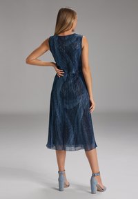 Femme aux longs cheveux blonds, portant une robe midi sans manches bleu marine et des sandales à talons hauts à lanières cheville bleu clair, se tenant avec une main sur la hanche.
