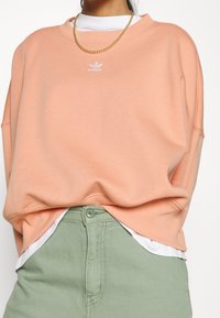 Persika oversized sweatshirt med ribbad rundhals och vitt logotyp på bröstet, bärs över en vit skjorta och matchas med ljusgröna byxor.