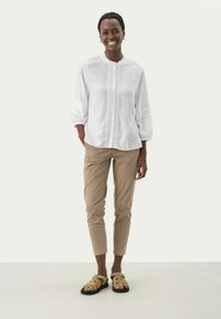 Witte linnen shirt met knopen aan de voorkant, gerimpelde kraag en plooien; beige broek; tan sandalen met uitsparingen en gespsluiting; model staat rechtop.