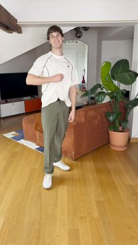 White t-shirt with black accents, olive green pants, and white sneakers; standing in a room with a potted plant and a textured orange couch.