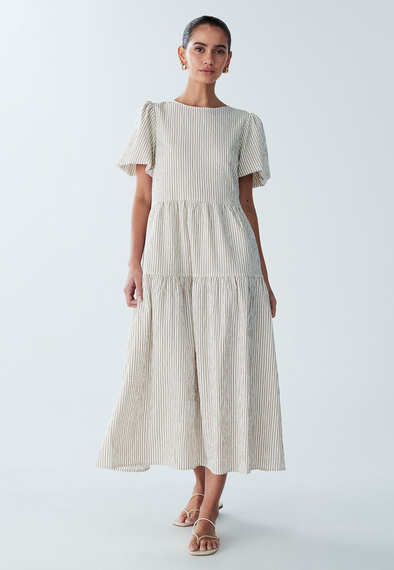 Robe midi rayée beige clair avec manches courtes, design à volants, tissu texturé et fines lignes horizontales. Portée avec des sandales neutres.