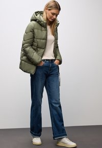 Kakigrön pufferjacka med huva, quiltad design, i kombination med blå vida jeans och vita sneakers, stående på en grå yta.