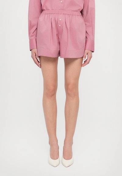 Shorts roses à taille élastique en tissu léger, dotés d'une silhouette décontractée et d'une texture lisse. Portés avec des talons hauts blancs.