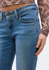 Κοντινό πλάνο προσώπου που φορά μπλε τζιν και καφέ καρ ώ μακρυμάνικη κορυφή κομμένη στη μέση, με το χέρι να ακουμπάει στον μηρό και ένα ασημένιο δαχτυλίδι στο δάχτυλο.