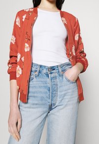 Veste bomber florale couleur rouille avec fermeture éclair, poignets côtelés et bande élastique blanche contrastante. Portée avec un jean en denim clair.