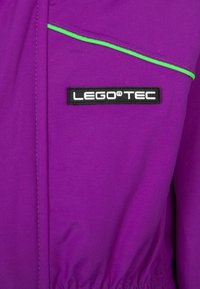 Fioletowa kurtka wykonana z trwałego materiału, z zielonym akcentem wzdłuż zamka błyskawicznego oraz czarną naszywką z logo oznaczoną "LEGO TEC". Elastyczny pas.