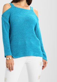 Pull en tricot turquoise avec un design décolleté, des manches longues et une texture distinctive. Associé à un jean blanc avec des accents en clous.
