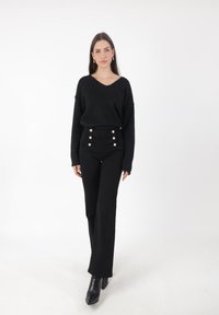 Kobieta stojąca w czarnym swetrze z dekoltem w kształcie litery V, czarnych spodniach o wysokim stanie z srebrnymi guzikami oraz czarnych botkach za kostkę na białym tle.