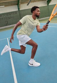 Kergelt roheline lühikeste varrukatega särk graafilise disainiga, valged tekstuuriga lühikesed püksid ja valged Nike tennisetossud sinisel tennisekursil.