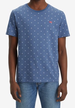 Homme portant un t-shirt bleu à manches courtes avec de petits motifs blancs en forme de losange et un logo Levi's rouge, assorti à un jean bleu.