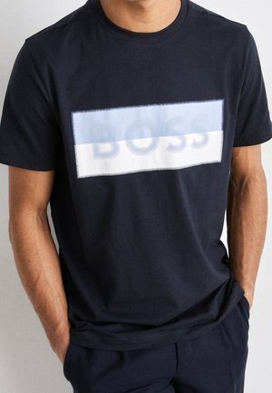 Homme portant un t-shirt bleu marine avec un logo rectangulaire bicolore bleu et blanc et le mot « BOSS » imprimé sur la poitrine.