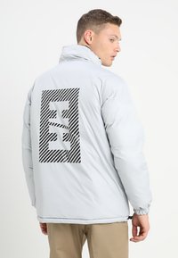 Šviesiai pilkas pūkinis švarkas su dideliu juodu dryžuotu logotipu ant nugaros, aukštu apykakle ir elastinėmis rankovėmis, užtikrinančiomis tvirtą prigludimą.