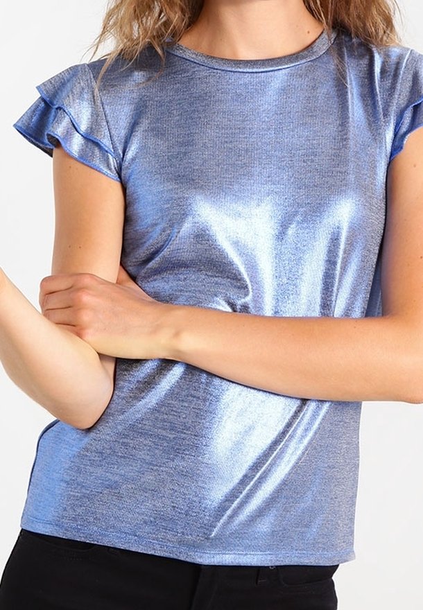 Femme portant un haut bleu métallique brillant à manches courtes avec des manches à volants, un pantalon noir, les bras croisés à la taille.