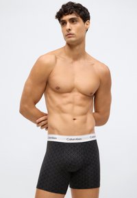 Černé boxerky vyrobené z měkké látky, s geometrickým vzorem, bílým pasem s textem "Calvin Klein" a přiléhavým střihem.