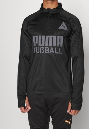 Musta pitkähihainen urheilu上o, jossa on neljänneksen vetoketju, teksturoitu kuvio ja suuri harmaa "PUMA FÜB3LL" -logo edessä.