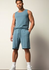 Lichtblauw tanktop en bijpassende korte broek van gestructureerde stof; de korte broek heeft een elastische tailleband en zijzakken. Witte sneakers maken de look compleet.