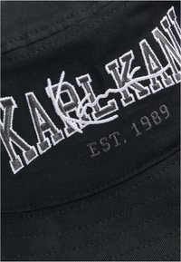 Sort bucket hat med broderet tekst "KARL KANI" og "EST. 1989" i hvid. Materialet har en glat tekstur med synlig syning.