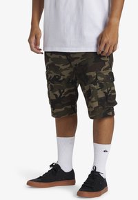 Camo cargoshorts in groen en bruin, met meerdere zakken. Draagt zwarte schoenen, witte sokken en een eenvoudige witte shirt.