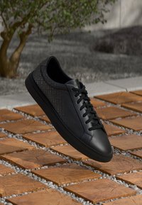 Sneaker in pelle nera con pannello laterale testurizzato, punta tondeggiante e chiusura con lacci, posizionato su uno sfondo di piastrelle in pietra.