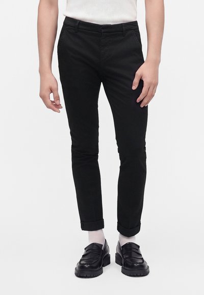 Personne vêtue d’un pantalon noir slim retroussé aux chevilles, de chaussettes blanches, de mocassins noirs et d’un top blanc sans manches, debout devant un fond uni.