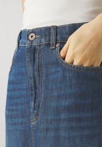 Jeans in denim blu a vita alta con una texture sbiadita, dotati di una tasca frontale visibile, bottone in metallo e dettagli di cucitura in contrasto.