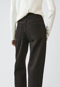 Bruine high-waisted jeans met een ontspannen pasvorm, voorzien van twee achterzakken en contrasterende stiksels, gecombineerd met een lichtgekleurde top met lange mouwen.