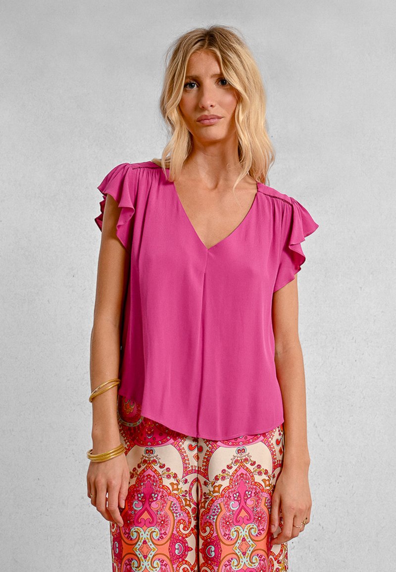 Blusa rosa a maniche corte con scollatura a V e maniche arricciate, abbinata a pantaloni a fantasia crema e rosa con un design floreale.
