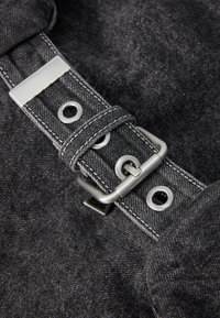 Tecido de denim preto com uma fivela de metal prateada e ilhós. Apresenta costura em contraste e uma superfície texturizada. Detalhe em aproximação.