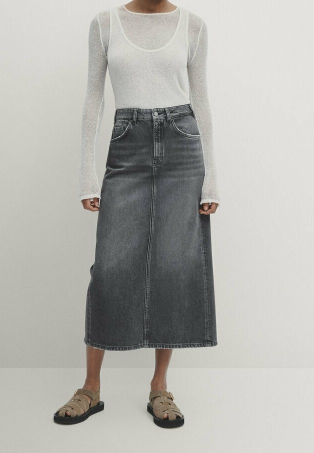 Femme portant un top blanc transparent à manches longues avec un débardeur blanc en dessous, une jupe midi en denim gris foncé taille haute et des sandales beiges.