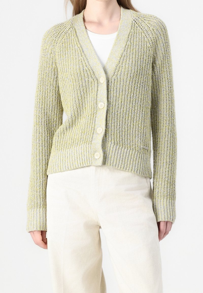 Cardigan tricoté texturé boutonné dans des tons vert clair et gris porté sur un haut blanc avec un pantalon couleur crème.