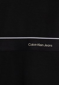 Czarna tkanina o gładkiej fakturze z białym poziomym paskiem i logo "Calvin Klein Jeans" w jasnym kroju na pasie.