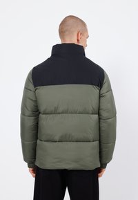 Chaqueta acolchada verde con sección superior negra, con costuras horizontales, puños elásticos y un cuello alto. Material suave y texturizado.