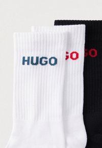 Βαμβακερές κάλτσες σε λευκό και μαύρο με ριγέ υφή. Η λευκή κάλτσα έχει μπλε λογότυπο «HUGO»· η μαύρη κάλτσα έχει κόκκινο λογότυπο «HUGO».