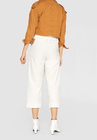 Brun cropped jakke med skulderepauleter parret med hvide cropped bukser og hvide højhælede ankelstøvler. Glat stof og struktureret pasform.