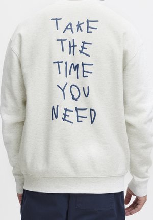 Personne portant un sweat-shirt gris clair avec un texte manuscrit bleu "Take the time you need" dans le dos et un pantalon foncé.