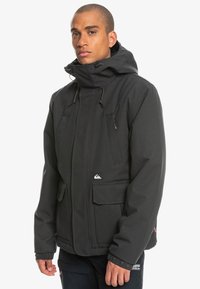 Schwarze Softshell-Jacke mit hohem Kragen, verstellbarem Kapuze, mehreren Fronttaschen und elastischen Bündchen. Mit einem Logo auf der unteren linken Seite.