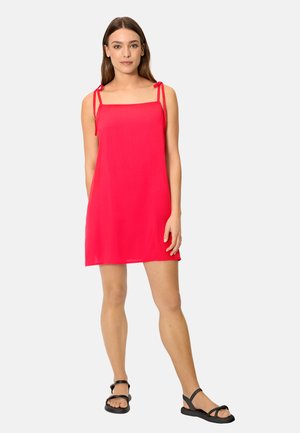 Mujer de pie que lleva un vestido mini sin mangas de color rojo brillante con tirantes atados en los hombros y sandalias planas negras sobre un fondo claro.