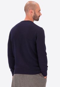 Uomo con barba ben curata che indossa un maglione a maniche lunghe blu scuro e pantaloni grigi, in piedi con le mani in tasca, girato di lato.