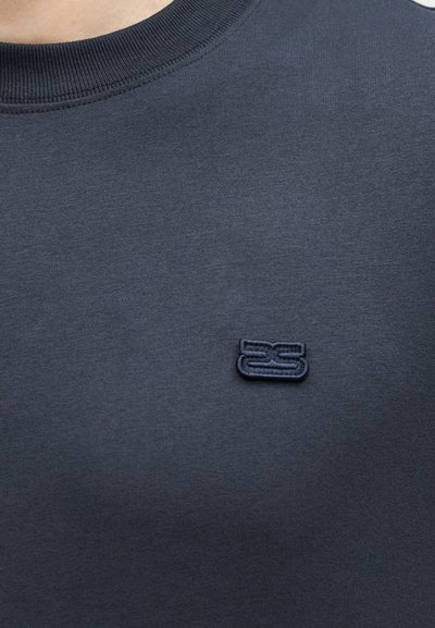 Κοντινό πλάνο μιας μπλε σκούρας μπλούζας με στρογγυλή λαιμόκοψη που διαθέτει ένα μικρό κεντημένο λογότυπο με στυλιζαρισμένα γράμματα στο στήθος.