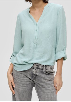 Frau trägt eine hellblaue, langärmelige Bluse mit aufgekrempelten Ärmeln, kombiniert mit ausgewaschenen grauen Jeans.