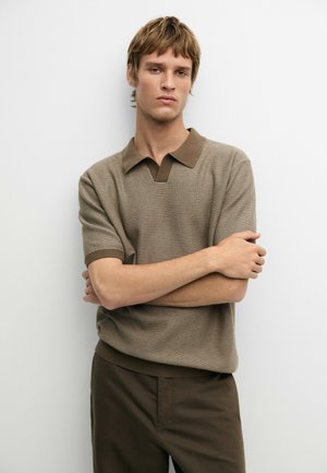 Giovane uomo con capelli castano chiaro indossa una polo a maniche corte in maglia e pantaloni marrone scuro, in piedi con le braccia incrociate su sfondo bianco.