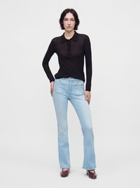 Femme aux cheveux courts et foncés portant une chemise noire transparente à manches longues, un jean bleu clair et des chaussures brun foncé, debout devant un fond blanc uni.