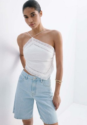 Femeie cu părul închis la culoare prins în spate, purtând un top alb asimetric din dantelă și pantaloni scurți din denim bleu, până la genunchi, cu brățări aurii la braț.