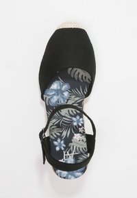 Černé plátěné espadrilky se zapínáním na kotník a modrým květinovým vzorem na stélce na bílém pozadí.