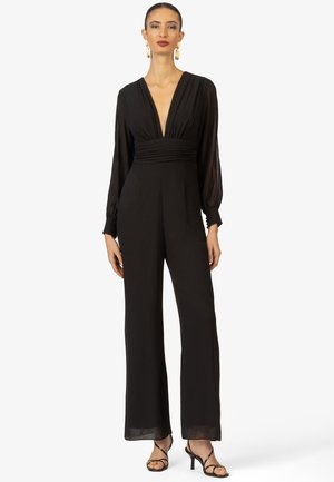 Zwarte jumpsuit met lange mouwen, diepe V-hals, geplooide taille en wijde benen. Doorzichtige stof bij de mouwen, gecombineerd met eenvoudige zwarte hakken.