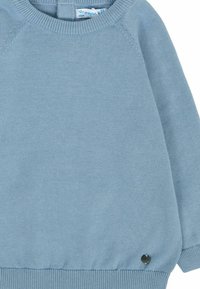 Pull en tricot bleu ciel avec un col rond côtelé, des manches raglan et un badge logo noir près de l'ourlet. Texture douce et coupe décontractée.