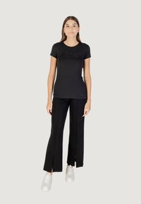Camisa negra de manga corta con un logo texturizado, combinada con pantalones negros con aberturas laterales, y con zapatillas blancas.