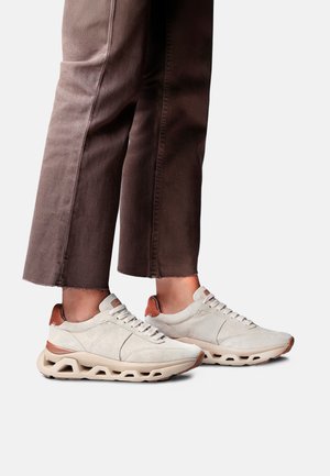 Personne portant des baskets beige clair et marron avec des semelles creuses uniques, associées à un pantalon marron court sur fond blanc.
