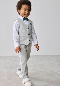 Gilet grigio chiaro con tre bottoni, camicia bianca, cravatta a farfalla blu, pantaloni grigi e sneakers bianche. Bambino che cammina con sicurezza.