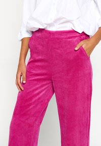 Pantaloni a gamba larga in velluto a coste rosa con una texture morbida, caratterizzati da una vita alta e tasche laterali, abbinati a una camicia bianca larga con bottoni.